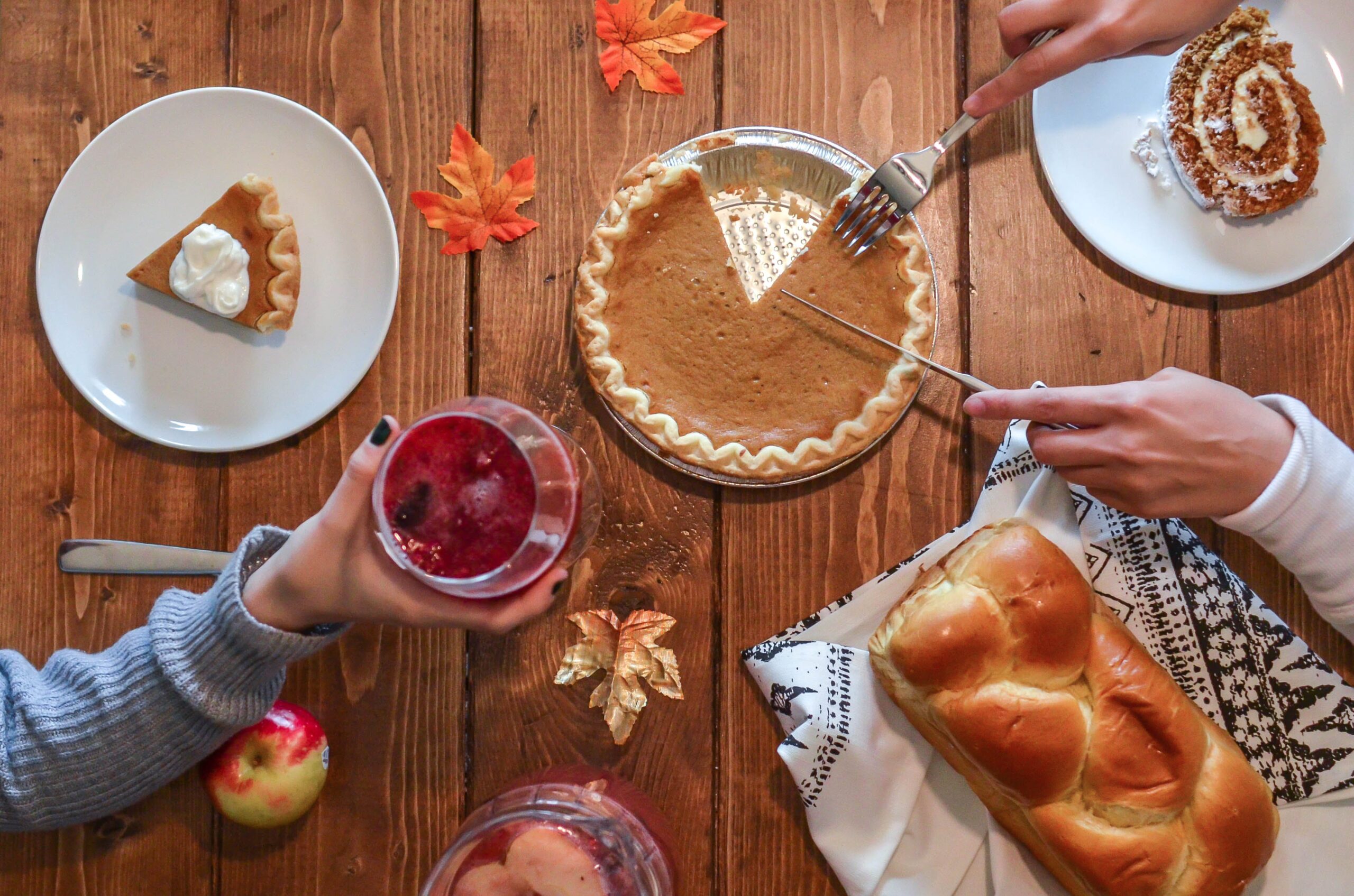 Someone's hands cut into a pumpkin pie with other Thanksgiving foods on the table
